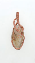 Load image into Gallery viewer, The #227 Cinnamon Montana Agate from Copper Den Stone Jewelry features a beige stone pendant intricately wrapped in oxidized copper wire swirls, creating an elegant loop at the top for wearing. The tightly coiled wire beautifully highlights the stone's smooth surface and natural pattern, enhancing its metaphysical properties.