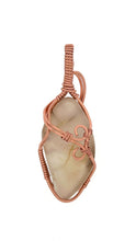 Load image into Gallery viewer, Product #227, the Cinnamon Montana Agate by Copper Den Stone Jewelry, features a polished beige stone encased in elaborate oxidized copper wirework. This pendant showcases decorative swirls and includes a loop at the top for hanging, highlighting its metaphysical properties against a plain white background.