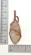 Load image into Gallery viewer, The #227 Cinnamon Montana Agate from Copper Den Stone Jewelry features a polished translucent stone wrapped in oxidized copper wire adorned with intricate swirls. When placed next to a ruler, it measures approximately 7 cm in length and 2.5 cm in width on a white background, highlighting its metaphysical properties.