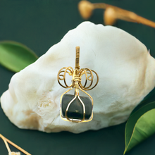 Load image into Gallery viewer, The #225 Shungite in Brass pendant by Copper Den Stone Jewelry, known for its EMF protection, showcases a dark gemstone wrapped in gold wire. It is displayed on a large, textured white shell surrounded by lush green leaves on a deep green surface.