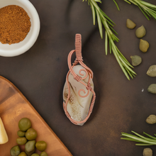 Load image into Gallery viewer, The #227 Cinnamon Montana Agate Pendant, handcrafted by Copper Den Stone Jewelry with intricate oxidized copper wire wrapping, is elegantly placed on a table. It is surrounded by green olives, capers, fresh rosemary sprigs, brown spices in a bowl, and the corner of a wooden board.